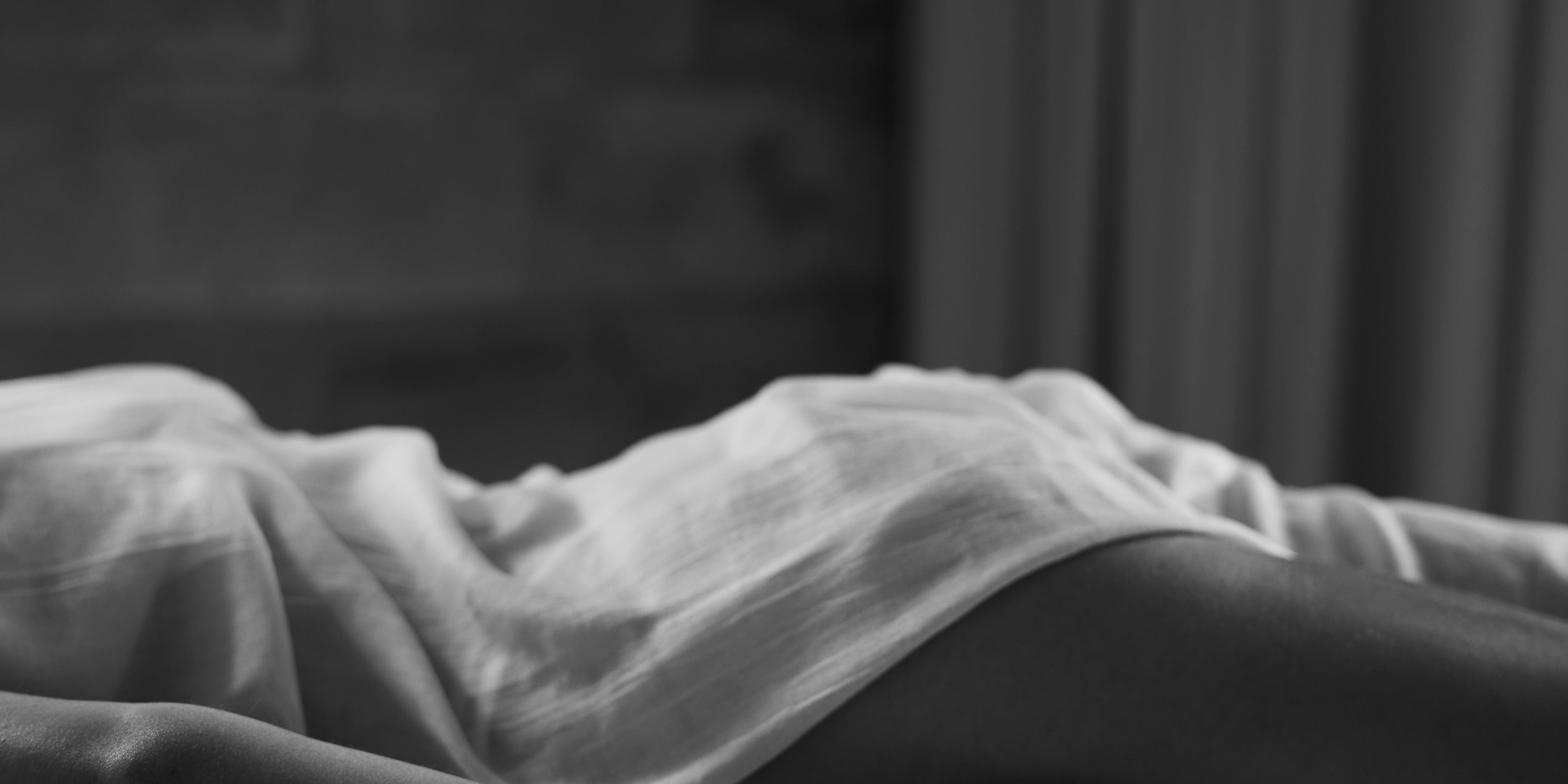 A person lies on a massage table, draped in a light sheet, with one arm relaxed by their side and palm open. The black and white image captures a calm, serene atmosphere with soft lighting and a spa-like setting.