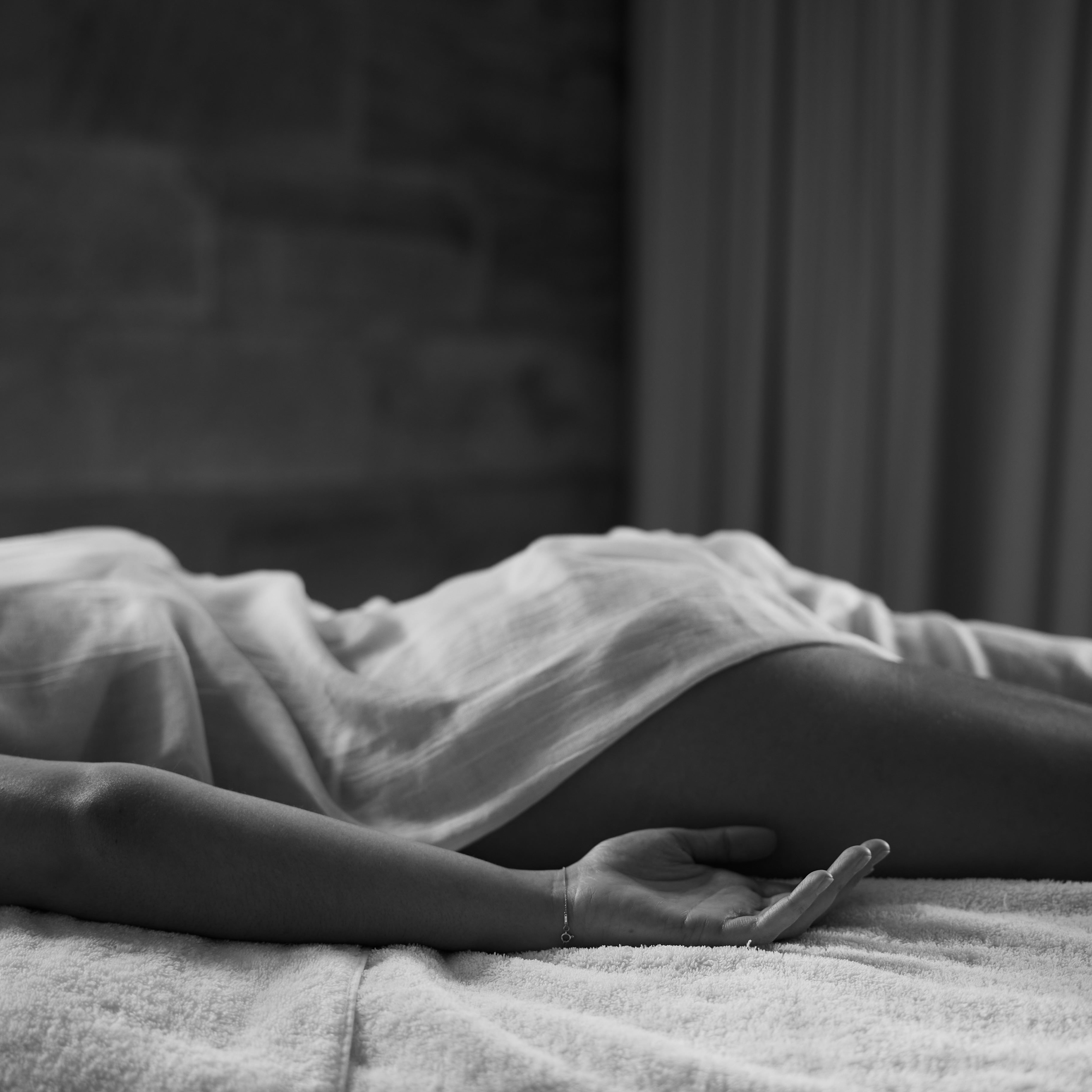 A person lies on a massage table, draped in a light sheet, with one arm relaxed by their side and palm open. The black and white image captures a calm, serene atmosphere with soft lighting and a spa-like setting.
