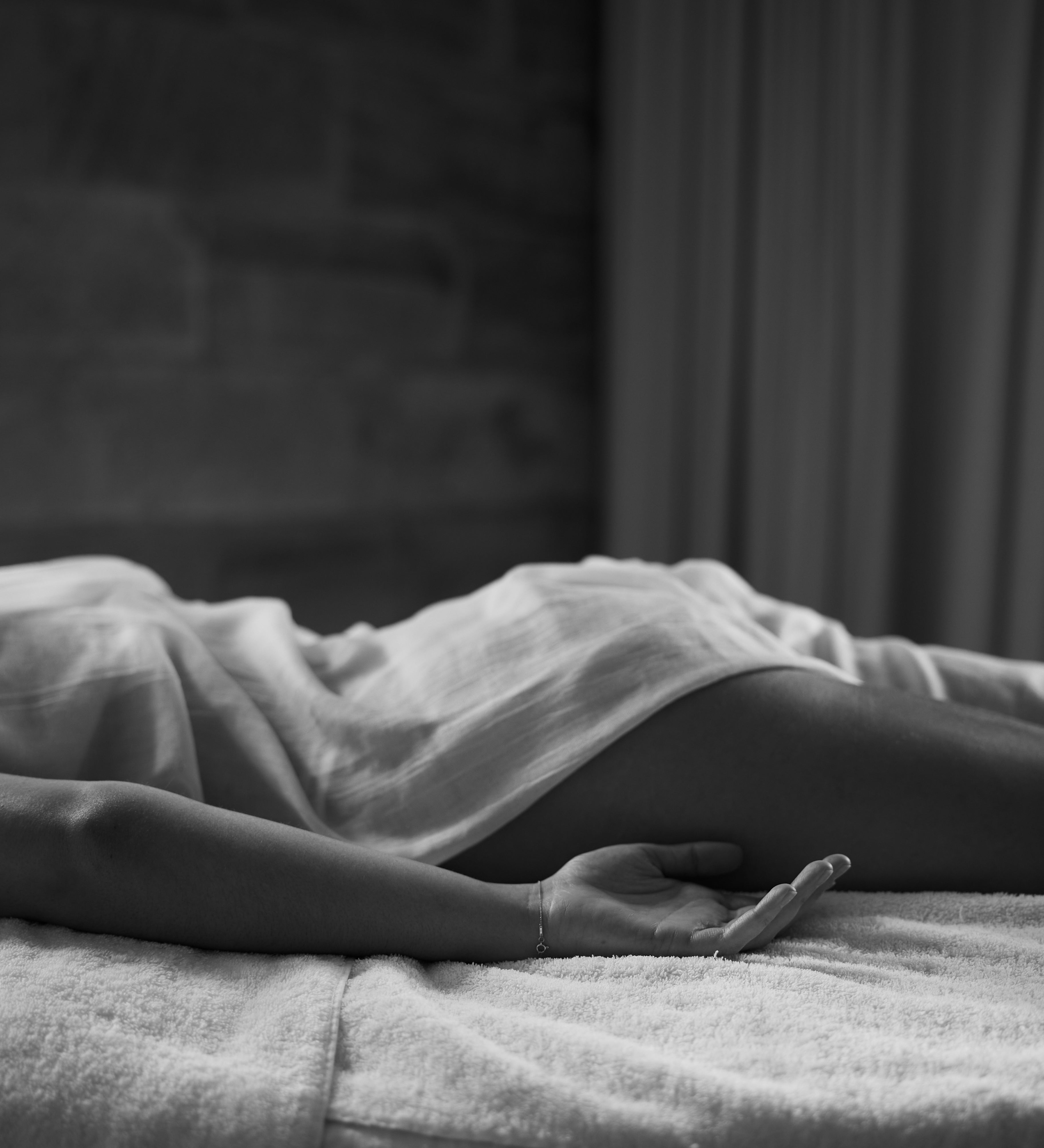 A person lies on a massage table, draped in a light sheet, with one arm relaxed by their side and palm open. The black and white image captures a calm, serene atmosphere with soft lighting and a spa-like setting.
