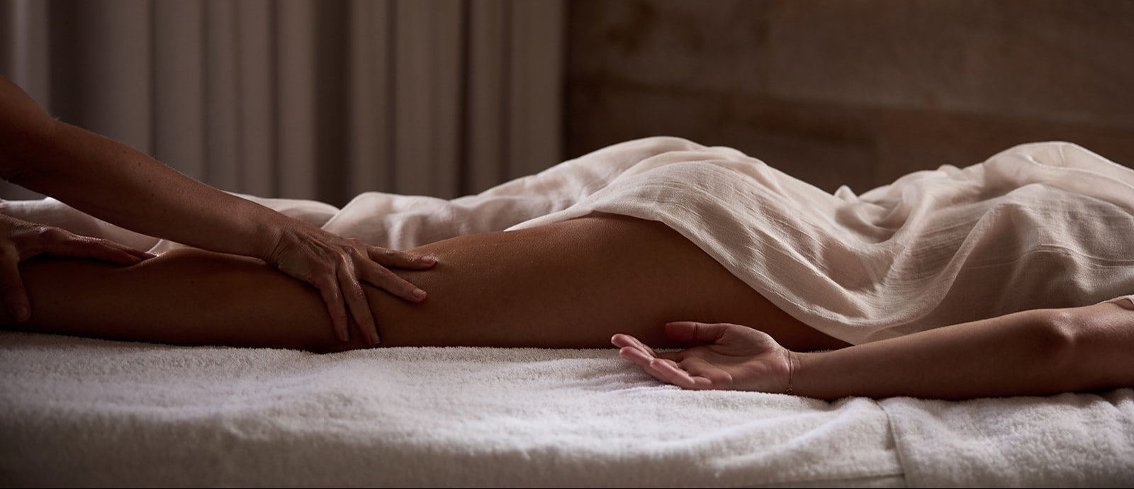 A person lies on a massage table covered with a sheer, neutral-toned cloth while receiving a relaxing leg massage in a softly lit spa setting. The calming ambiance is enhanced by warm tones and minimal background distractions.